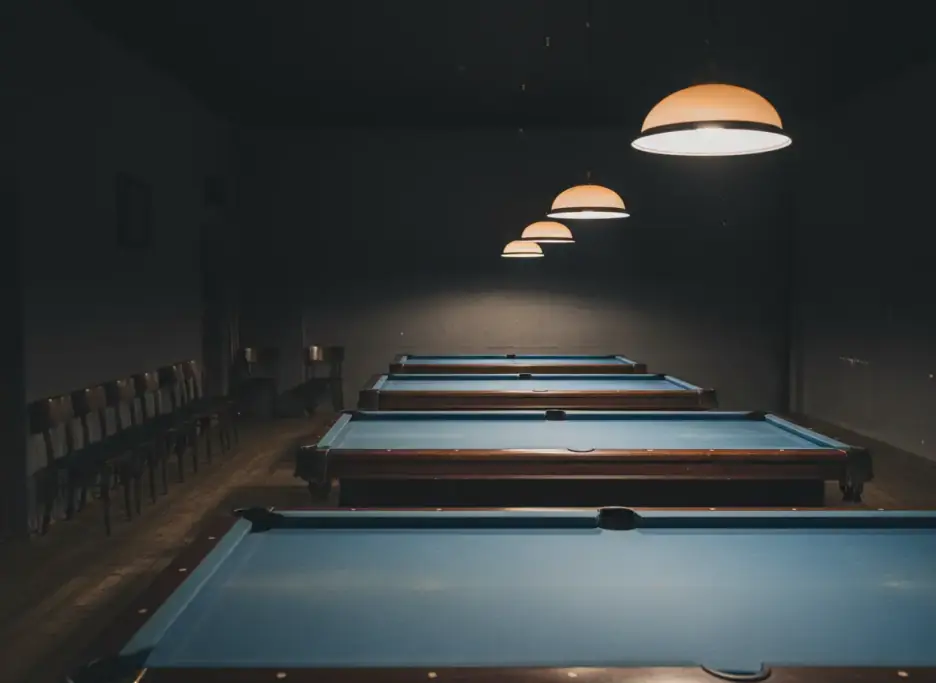 A row of blue-cloth pool tables under hanging lamps in an empty billiards room